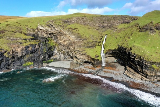 Aerial view of Waterstein Head with a waterfall cascading down dramatic cliffs onto a rocky beach along the coastline of the Isle of Skye Scotland United Kingdom. Aerial view of Waterstein Head with a waterfall cascading down dramatic cliffs onto a rocky beach along the coastline of the Isle of Skye Scotland United Kingdom.