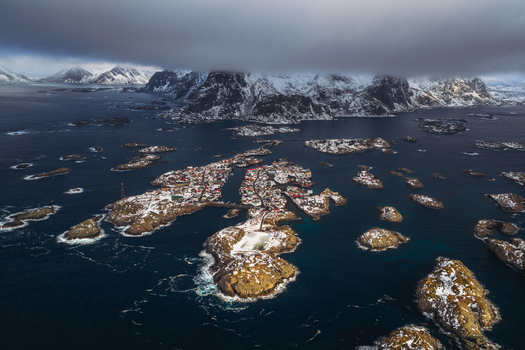 Aerial view of a quaint village nestled amid rugged snow dusted islands surrounded by the deep blue sea under a blanket of clouds Henningsv&aelig;r Nordland Norway.