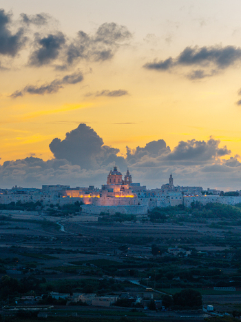 Aerial view of the fortified city of Mdina illuminated at sunset with golden light on its stone walls and cathedral dome surrounded by rural landscape in Mdina Western Region Malta.
