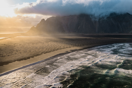 Aerial view of Vestrahorn mountain rising above the black sand beach and crashing ocean waves under a dramatic cloudy sky during golden hour in Sveitarfelagid Hornafjordur Iceland. Aerial view of Vestrahorn mountain rising above the black sand beach and crashing ocean waves under a dramatic cloudy sky during golden hour in Sveitarfelagid Hornafjordur Iceland.