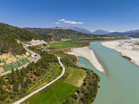 Aerial view of Vjosa Wild River National Park with its winding turquoise river lush green fields and a road running alongside the mountains in Permet Gjirokaster County Albania. Aerial view of Vjosa Wild River National Park with its winding turquoise river lush green fields and a road running alongside the mountains in Permet Gjirokaster County Albania.