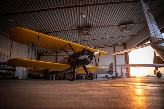 View of a vintage Boeing Stearman biplane with yellow wings and a dark blue fuselage parked inside a hangar at sunset in Langenhagen Niedersachsen Germany. View of a vintage Boeing Stearman biplane with yellow wings and a dark blue fuselage parked inside a hangar at sunset in Langenhagen Niedersachsen Germany.