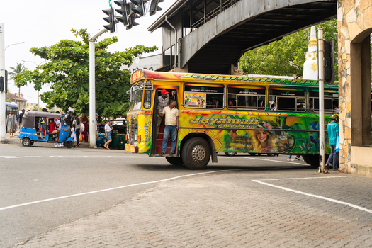 Sri Lanka 07 January 2026 View of a colorful private bus stopped at a busy intersection under a pedestrian bridge with a blue tuk tuk and pedestrians nearby.