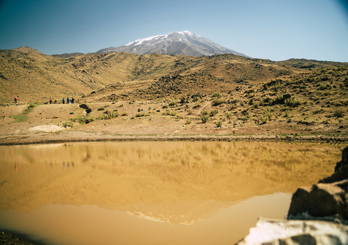 View of Mount Ararat reflecting in a muddy pond with a group of people walking on the rocky hillside in Turkey.