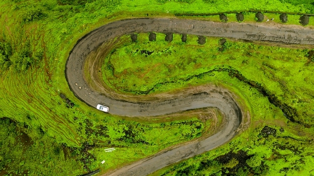 Aerial view of a winding S shaped road cutting through lush green hills with a white car driving along the curve in Igatpuri Maharashtra India. Aerial view of a winding S shaped road cutting through lush green hills with a white car driving along the curve in Igatpuri Maharashtra India.
