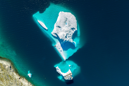 Aerial view of a large iceberg floating in deep blue water with turquoise glacial meltwater surrounding the ice near a rocky coastline in Greenland. Aerial view of a large iceberg floating in deep blue water with turquoise glacial meltwater surrounding the ice near a rocky coastline in Greenland.