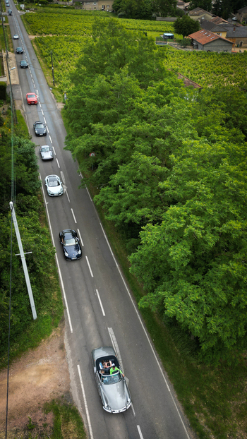 Aerial view of a convoy of sports cars driving along a rural road bordered by lush green trees and vineyards La Chapelle de Guinchay Bourgogne Franche Comte France.