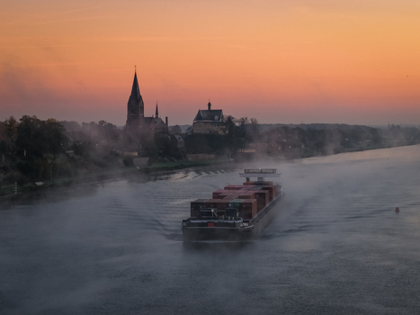 Aerial view of a container barge navigating the misty Meuse river at sunrise with the silhouette of Kessel castle and church spire in the background Kessel Limburg Netherlands.