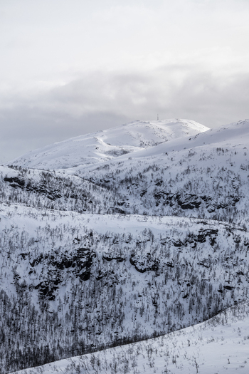 View of snow covered mountain peaks under a cloudy sky with sparse vegetation on the slopes in Ersfjord Troms Norway. View of snow covered mountain peaks under a cloudy sky with sparse vegetation on the slopes in Ersfjord Troms Norway.
