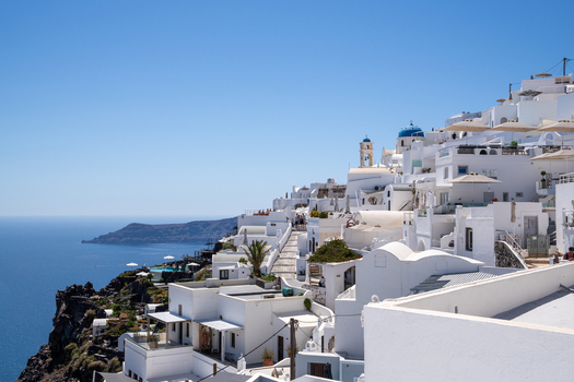 View of white houses and blue domed churches on the volcanic cliffs of Santorini overlooking the deep blue Aegean Sea under a clear sky in Fira Greece. View of white houses and blue domed churches on the volcanic cliffs of Santorini overlooking the deep blue Aegean Sea under a clear sky in Fira Greece.