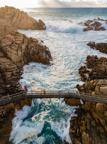 Aerial view of Canal Rocks Bridge with a person walking across the wooden footbridge over crashing ocean waves and rugged granite rocks in Yallingup Western Australia Australia. Aerial view of Canal Rocks Bridge with a person walking across the wooden footbridge over crashing ocean waves and rugged granite rocks in Yallingup Western Australia Australia.