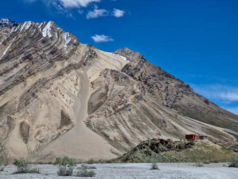 View of the rugged mountainous landscape along the Penzi La road with rocky slopes sparse vegetation and a small building on a hill under a clear blue sky in Zanskar Ladakh India.