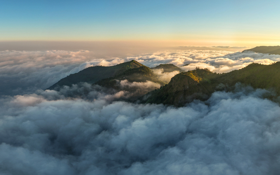 Aerial view of Dieng Plateau peaks rising above a dense sea of clouds during golden hour in Indonesia.
