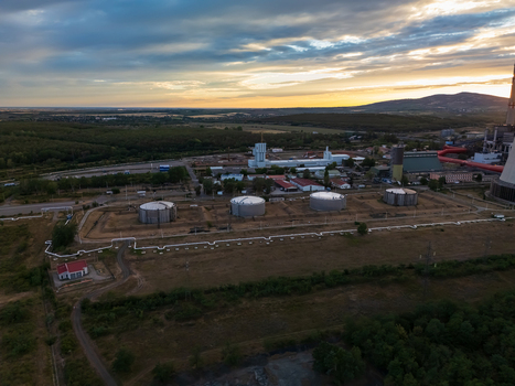 Aerial view of the Matra power plant storage tanks pipelines and industrial buildings beside wooded farmland and rolling hills at sunset in Visonta Hungary.