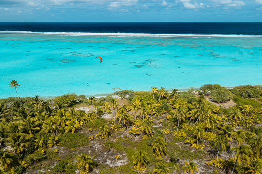 Aerial view of Aitutaki lagoon with lush palm trees and a kitesurfer gliding over turquoise water in Cook Islands. Aerial view of Aitutaki lagoon with lush palm trees and a kitesurfer gliding over turquoise water in Cook Islands.