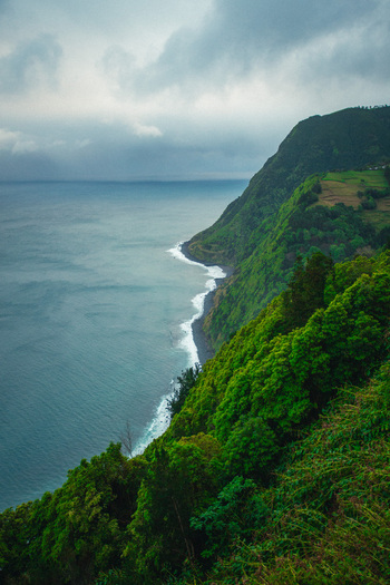 Aerial view of the dramatic coastline where verdant cliffs meet the dark swirling ocean under a brooding sky Nordeste Azores Portugal.