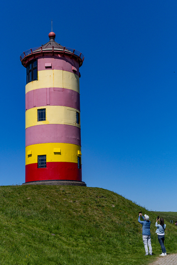 View of the colorful Pilsum lighthouse with its iconic red yellow and pink stripes on a green dike as tourists take photos under a clear blue sky Krummhorn Niedersachsen Germany. View of the colorful Pilsum lighthouse with its iconic red yellow and pink stripes on a green dike as tourists take photos under a clear blue sky Krummhorn Niedersachsen Germany.