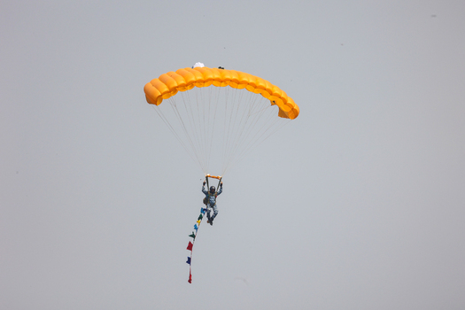 Dhaka Bangladesh 25 March 2026 View of a lone skydiver suspended beneath a vibrant orange parachute trailing colorful flags against the muted sky.
