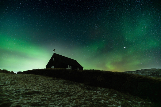 View of a dark church silhouette atop a hill against a vibrant green aurora borealis dancing across a starry night sky Krysuvikurkirkja Hafnarfjardarkaupstadur Iceland.