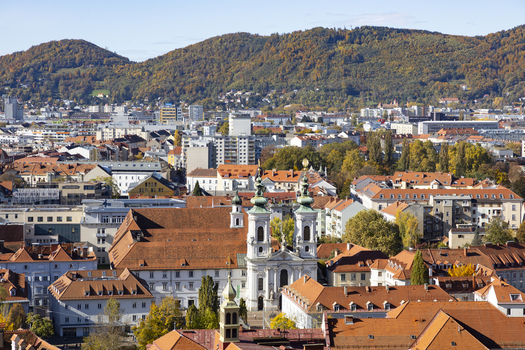 View of white church with green dome under a clear blue sky surrounded by buildings with orange roofs and mountains in the background Graz Styria Austria. View of white church with green dome under a clear blue sky surrounded by buildings with orange roofs and mountains in the background Graz Styria Austria.