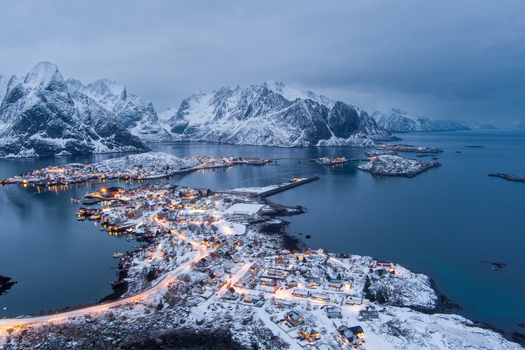 Aerial view of the snow covered village of Lofoten with illuminated houses winding roads and dramatic mountains reflecting in the calm sea in Nordland Norway.