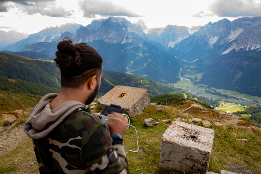 View of a man operating a drone remote controller on a mountain summit overlooking the jagged peaks of the Dolomites and a lush green valley Rocca Pietore Veneto Italy. View of a man operating a drone remote controller on a mountain summit overlooking the jagged peaks of the Dolomites and a lush green valley Rocca Pietore Veneto Italy.