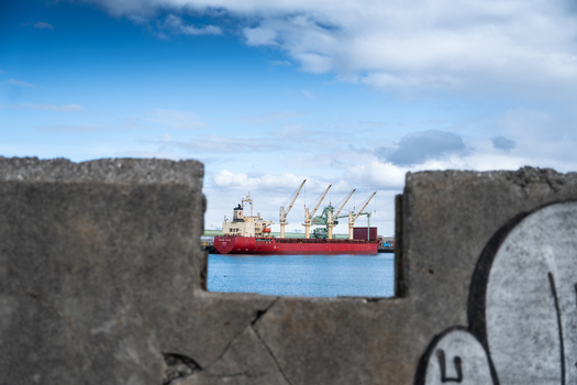 Hafnarfjordur Iceland 17 April 2026 View of a large red cargo ship docked at the port with cranes visible through a gap in a concrete wall under a cloudy sky.