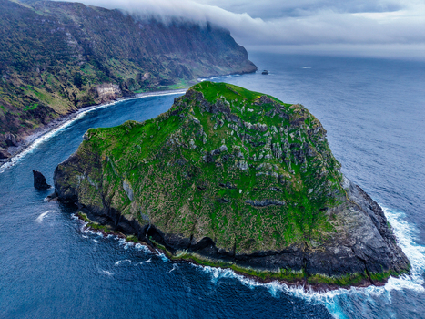 Aerial view of a verdant island contrasting with the dark blue sea under a misty sky near Estrada do Farol Flores A&ccedil;ores Portugal.