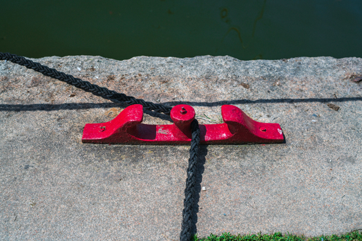 View of a red metal mooring cleat with a black rope on a stone quay in Charlestown Cornwall United Kingdom. View of a red metal mooring cleat with a black rope on a stone quay in Charlestown Cornwall United Kingdom.