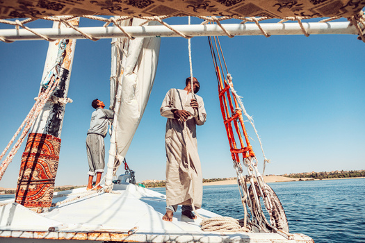View of two men operating a traditional felucca sailboat on the Nile River with desert hills in the background under a clear blue sky Aswan Aswan Governorate Egypt. View of two men operating a traditional felucca sailboat on the Nile River with desert hills in the background under a clear blue sky Aswan Aswan Governorate Egypt.