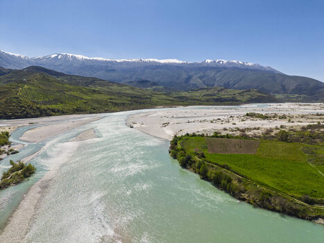 Aerial view of Vjosa Wild River National Park with turquoise river gravel banks green fields and snow capped mountains under a clear blue sky in Permet Gjirokaster County Albania. Aerial view of Vjosa Wild River National Park with turquoise river gravel banks green fields and snow capped mountains under a clear blue sky in Permet Gjirokaster County Albania.