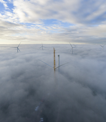 Aerial view of wind turbines emerging from a thick layer of white fog with a yellow construction crane under a blue sky in Netherlands. Aerial view of wind turbines emerging from a thick layer of white fog with a yellow construction crane under a blue sky in Netherlands.