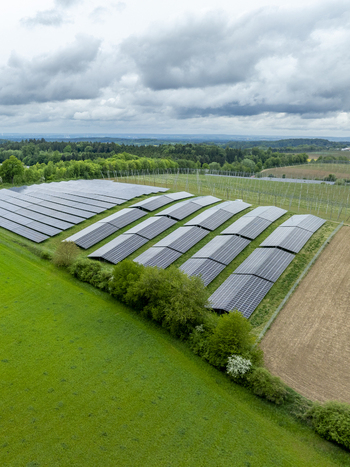 Aerial view of a solar panel farm surrounded by lush green fields and dense forests under a cloudy sky in Aiglsbach Bavaria Germany.