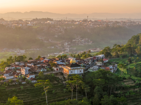 Aerial view of a village with a mosque featuring a golden dome surrounded by lush terraced fields and misty mountains during golden hour in Indonesia.