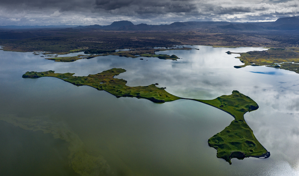 Aerial view of the green pseudocraters of Lake Myvatn surrounded by calm waters and distant volcanic mountains under a cloudy sky in Iceland.