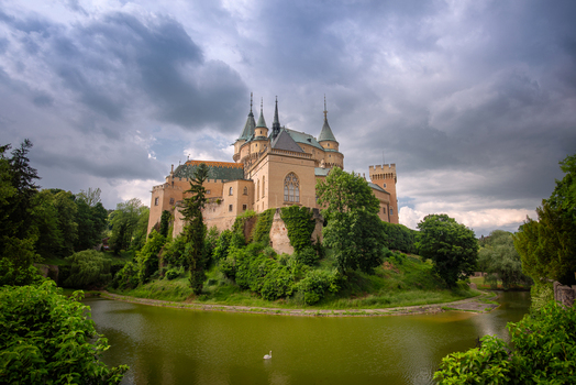 View of Bojnice Castle with its medieval towers and stone walls rising above a green moat surrounded by lush trees under a dramatic cloudy sky in Prievidza Trenciansky kraj Slovakia.
