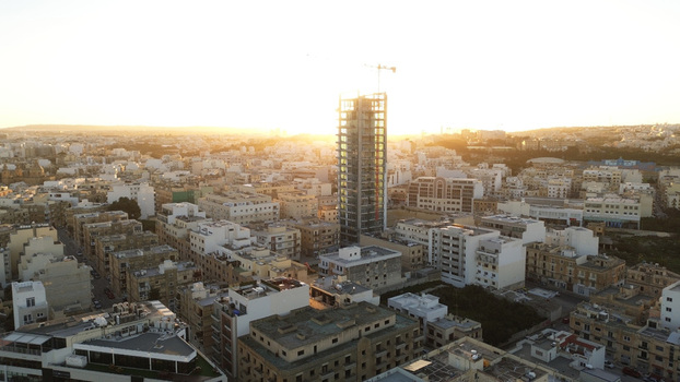 Aerial view of a modern high rise building under construction rising above the dense urban landscape during golden hour with warm sunlight in Valletta Port Region Malta. Aerial view of a modern high rise building under construction rising above the dense urban landscape during golden hour with warm sunlight in Valletta Port Region Malta.