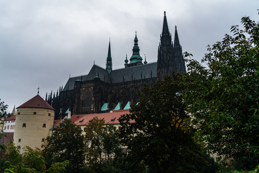 View of the historic St. Vitus Cathedral with its towering Gothic spires rising above stone fortifications and lush trees in Prague Prague Czechia.