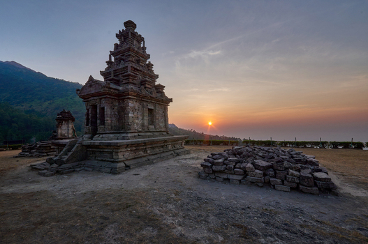 View of the ancient Gedong Songo Temple complex with stone ruins and a mountain backdrop during an orange sunset in Bandungan Jawa Tengah Indonesia.