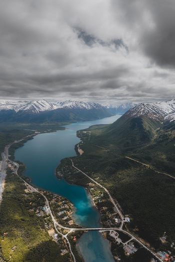 Aerial view of Alaska Wilderness featuring a deep blue lake surrounded by snow capped mountains dense forests and a winding road in Palmer Alaska United States. Aerial view of Alaska Wilderness featuring a deep blue lake surrounded by snow capped mountains dense forests and a winding road in Palmer Alaska United States.