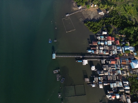 Aerial view of Galang Baru coastal village with traditional houses on stilts a long wooden pier and floating fish farms along the shore in Galang Riau Islands Indonesia. Aerial view of Galang Baru coastal village with traditional houses on stilts a long wooden pier and floating fish farms along the shore in Galang Riau Islands Indonesia.