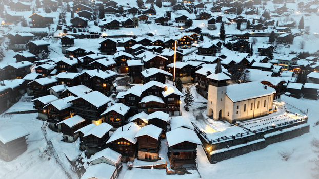 Aerial view of snow dusted chalets nestled around a brightly lit church creating a warm glow against the cool crisp winter landscape Grimentz Switzerland.