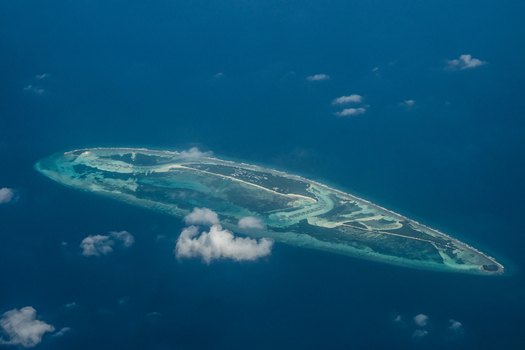 Aerial view of an island surrounded by turquoise waters and scattered clouds creating a serene contrast between the land and sea Flight from Male to Hanimaadhoo island Maldives.
