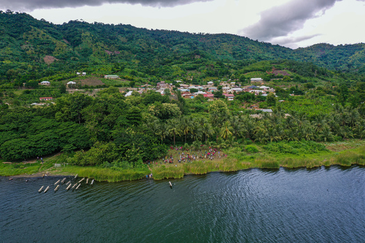 Aerial view of Lake Bosomtwe with a village nestled among lush greenery traditional wooden boats moored along the shore and people gathered near the water in Bosomtwe Ashanti Region Ghana.