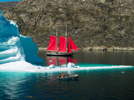 Aerial view of a sailboat with vibrant red sails near a massive iceberg in icy waters with a small inflatable boat nearby and rugged rocky mountains in the background Greenland. Aerial view of a sailboat with vibrant red sails near a massive iceberg in icy waters with a small inflatable boat nearby and rugged rocky mountains in the background Greenland.