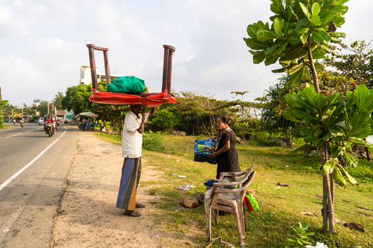 Sri Lanka 15 January 2026 View of a man carrying a wooden table on his head while a woman stands nearby on a roadside with lush green trees and a cloudy sky.