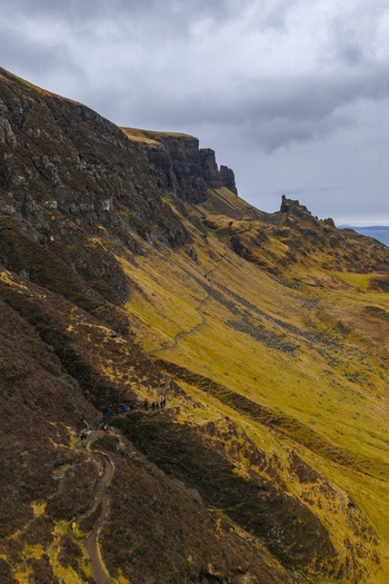 Aerial view of The Quiraing landslip with hikers on a winding trail along steep grassy slopes under a cloudy sky Isle of Skye Scotland United Kingdom. Aerial view of The Quiraing landslip with hikers on a winding trail along steep grassy slopes under a cloudy sky Isle of Skye Scotland United Kingdom.