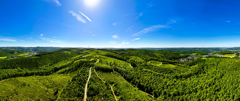 Aerial view of a wide panorama featuring rolling green hills and dense forests under a bright sun and clear blue sky near Siegen North Rhine Westphalia Germany.