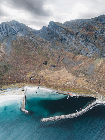 Aerial view of the rugged coastline and harbor in Senja featuring stone breakwaters turquoise waters and dramatic mountains under a cloudy sky in Senja Troms Norway.
