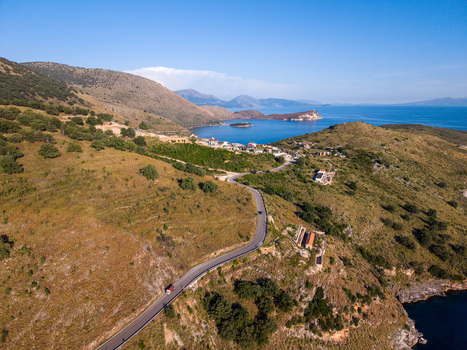 Aerial view of a winding asphalt road on a grassy hillside overlooking the turquoise waters of the bay and the historic fortress Porto Palermo Qarku i Vlores Albania. Aerial view of a winding asphalt road on a grassy hillside overlooking the turquoise waters of the bay and the historic fortress Porto Palermo Qarku i Vlores Albania.